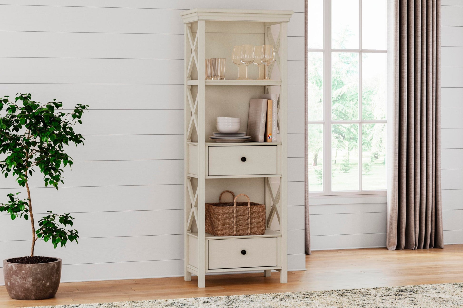 The Bolanburg Display Cabinet in antiqued white stands by a potted plant and window on wood floors, showcasing two drawers, glassware, books, and a basket for charming cottage-style appeal in a bright room.