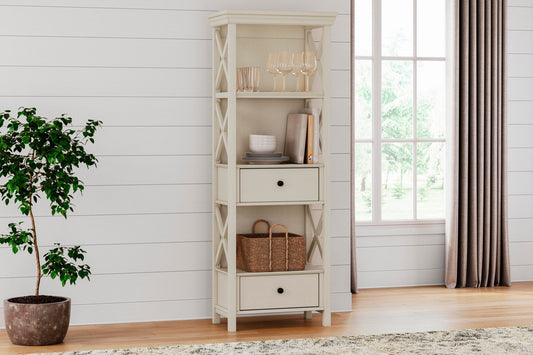 The Bolanburg Display Cabinet in antiqued white stands by a potted plant and window on wood floors, showcasing two drawers, glassware, books, and a basket for charming cottage-style appeal in a bright room.