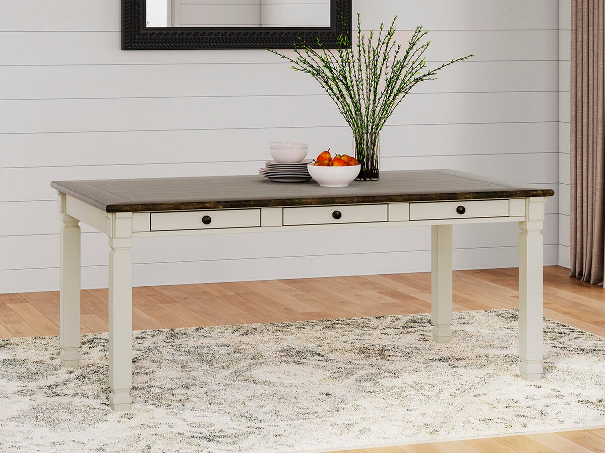 The Bolanburg Rectangular Dining Room Table with white legs and a dark top sits on a light patterned rug, decorated with a vase of branches, bowls, and a plate of fruit.