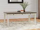 The Bolanburg Rectangular Dining Room Table with white legs and a dark top sits on a light patterned rug, decorated with a vase of branches, bowls, and a plate of fruit.