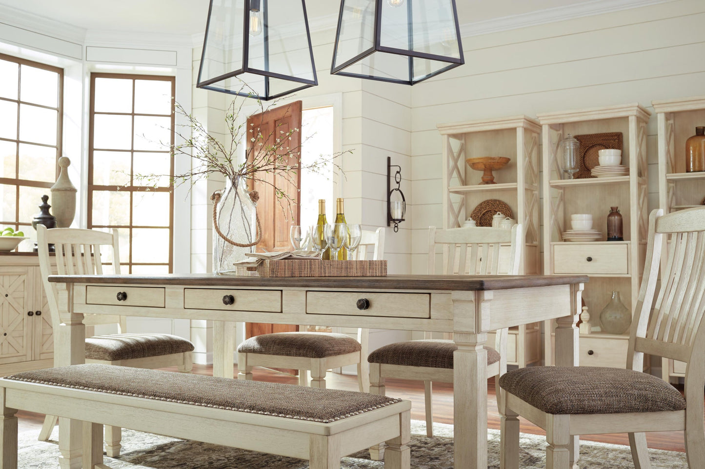 A bright dining room features the Bolanburg Rectangular Dining Room Table with six chairs, a bench, pendant lights, shelves displaying decor and dishes, plus a vase with branches and wine bottles on the table.