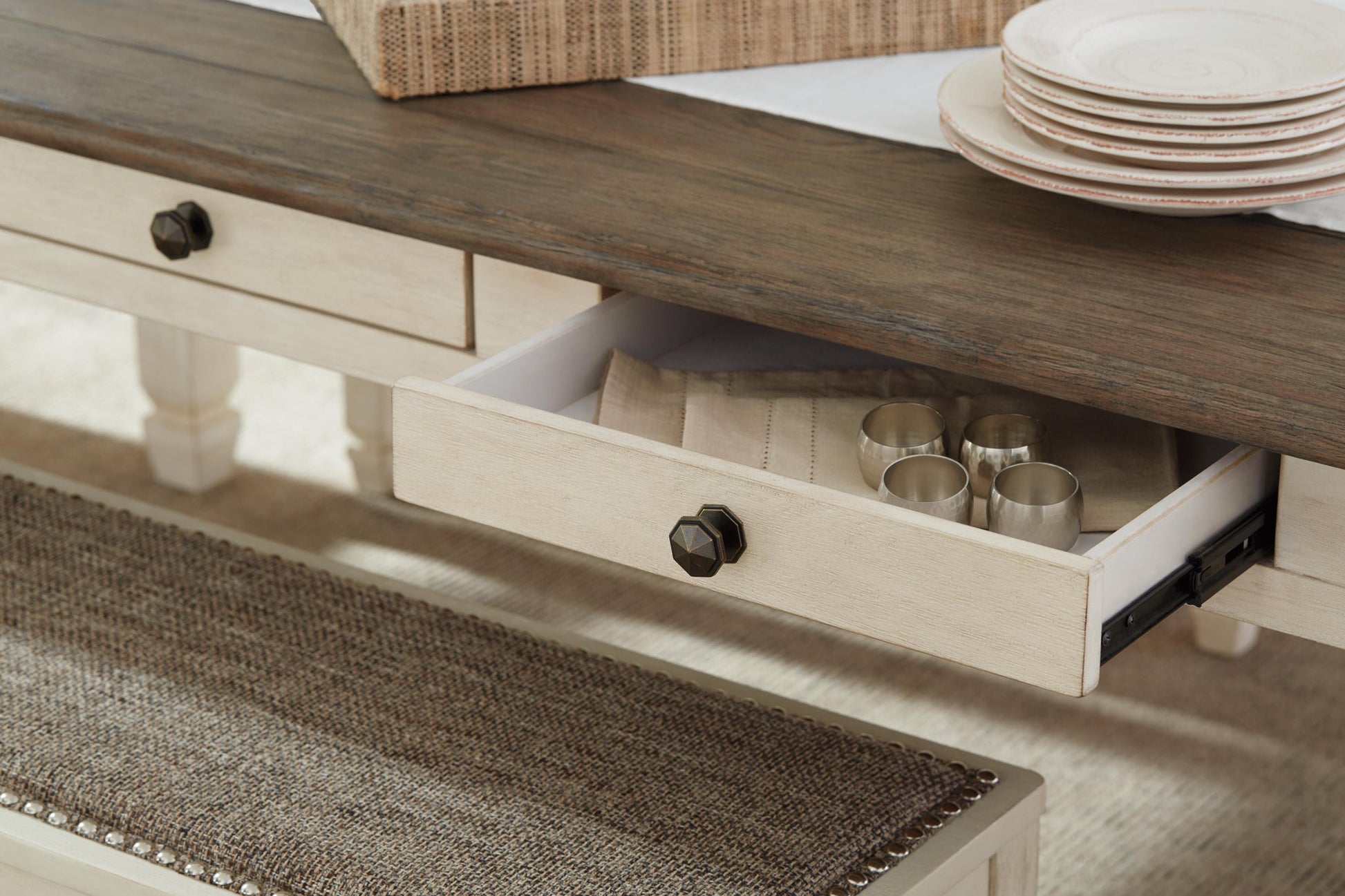A close-up of the Bolanburg Rectangular Dining Room Table shows an open drawer with napkins and four metal cups, while stacked plates and a woven tray sit atop the elegant two-tone table.