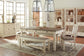 A dining room featuring the Bolanburg Rectangular Dining Room Table with drawers, four chairs, a bench, light cabinets, glass decor, and two geometric pendant lights on a patterned rug.