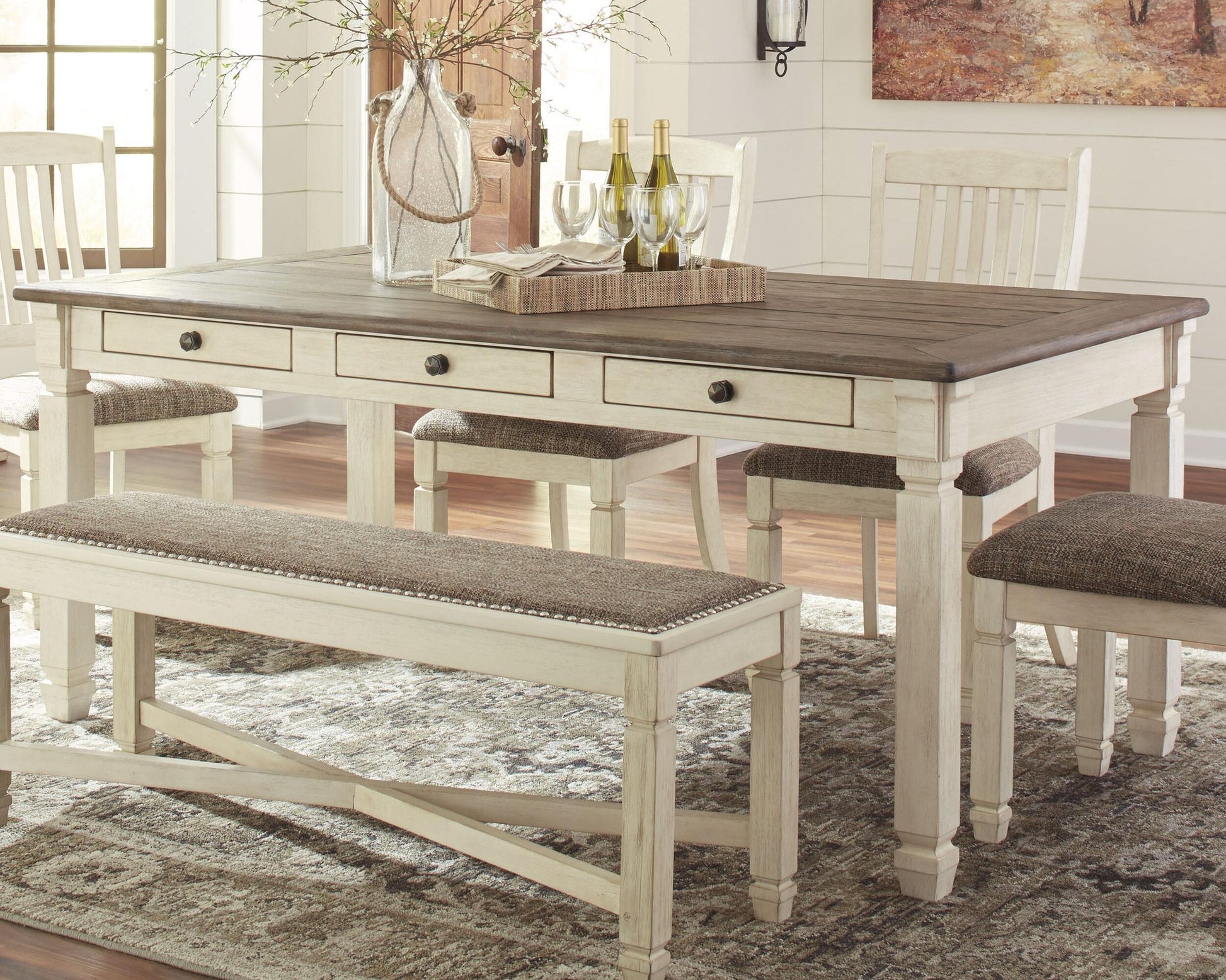 The Bolanburg Rectangular Dining Room Table is paired with four chairs and a bench, styled with a wooden tray holding wine bottles and glasses, and positioned on a patterned rug in a bright room.