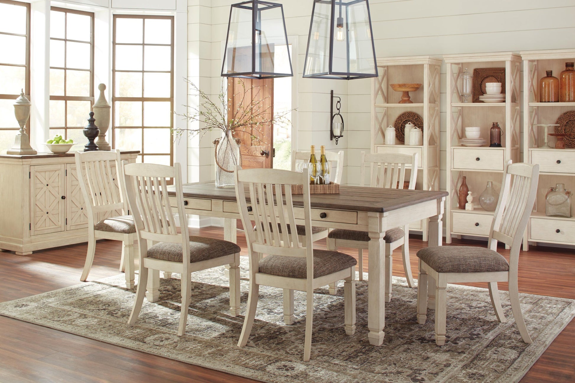 Bright dining room with a Bolanburg Rectangular Dining Room Table, six chairs, two pendant lights, and shelves displaying decorative items on a patterned rug.