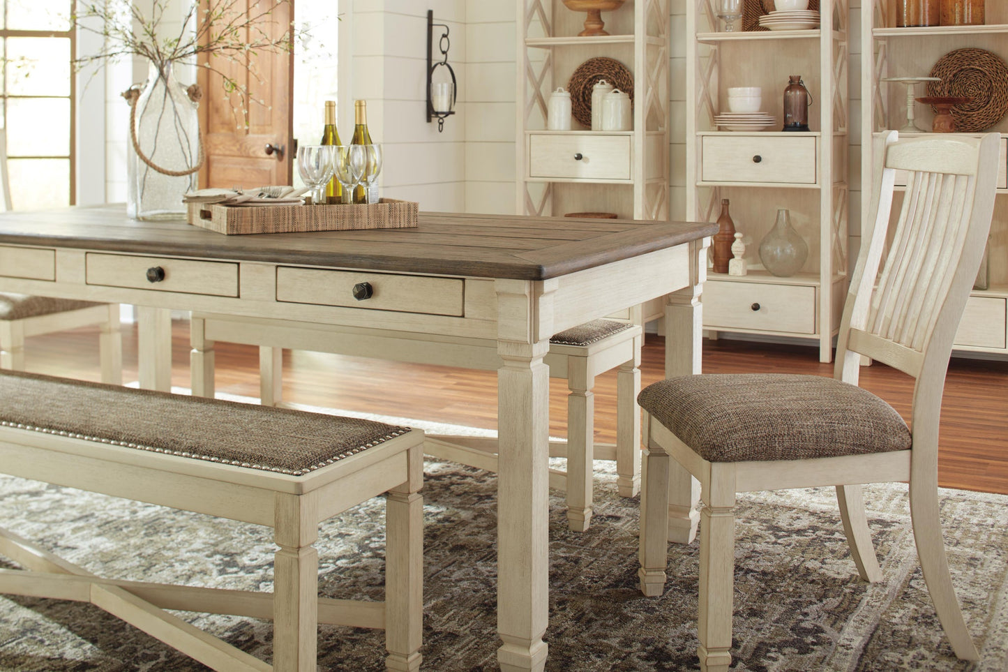 A dining room featuring the Bolanburg Rectangular Dining Room Table, a matching bench and chair, a decorative tray with bottles and glasses, and shelves in the background displaying ceramics and vases.