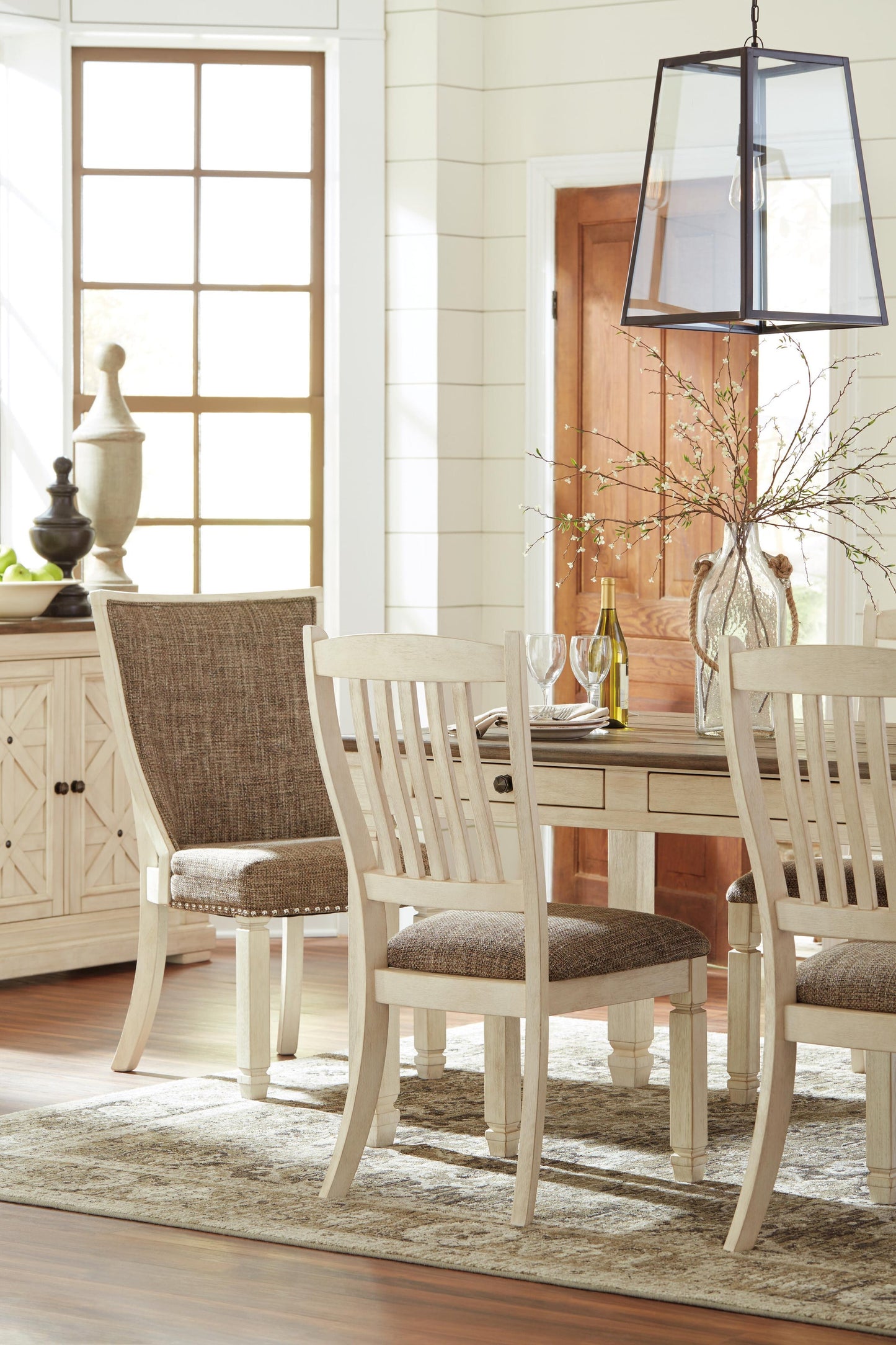 Bright dining room featuring the Bolanburg Rectangular Dining Room Table, four upholstered chairs, a glass vase with branches, a wine bottle, and a large window that fills the space with natural light.