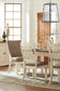 Bright dining room featuring the Bolanburg Rectangular Dining Room Table, four upholstered chairs, a glass vase with branches, a wine bottle, and a large window that fills the space with natural light.