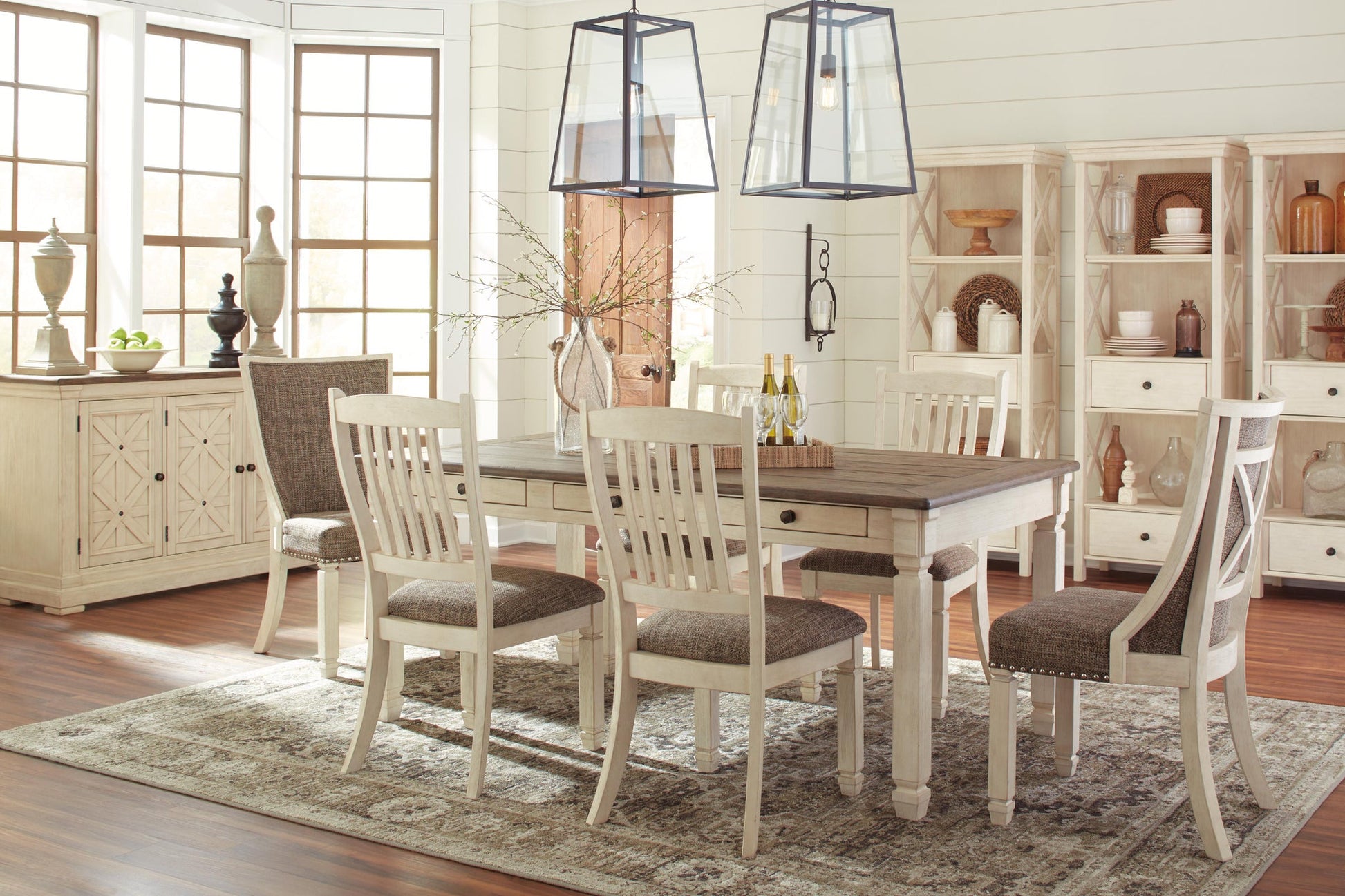 A dining room featuring a wooden table, four Bolanburg Dining UPH Side Chairs with cushioned seats, a sideboard, decorative shelves, and two pendant lights— all in neutral tones with sunlight streaming through large windows.