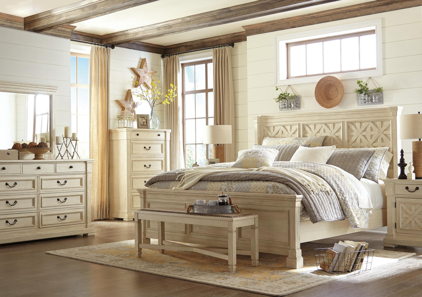Bright bedroom featuring the Bolanburg Large UPH Dining Room Bench in an antiqued white finish, cream-colored wooden furniture, a large bed with patterned bedding, dresser, chest, beige curtains, and decorative wall accents.