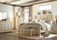 Bright bedroom featuring the Bolanburg Large UPH Dining Room Bench in an antiqued white finish, cream-colored wooden furniture, a large bed with patterned bedding, dresser, chest, beige curtains, and decorative wall accents.