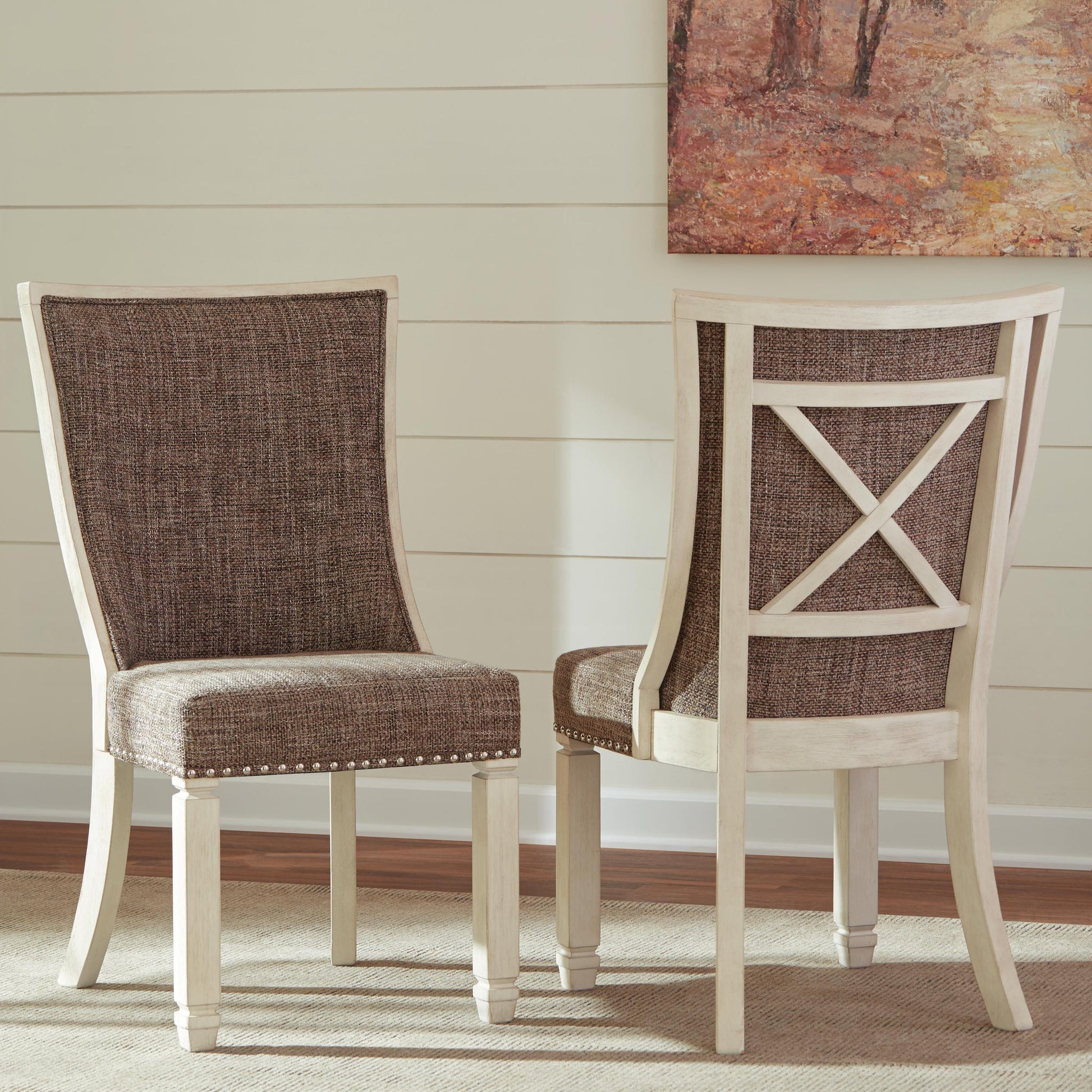 Two Bolanburg Dining UPH Side Chairs (2/CN) with antiqued white wooden frames shown—one facing forward, the other backward to display an X-shaped backrest. The setting also features a painting and white wall paneling.
