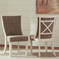 Two Bolanburg Dining UPH Side Chairs (2/CN) with antiqued white wooden frames shown—one facing forward, the other backward to display an X-shaped backrest. The setting also features a painting and white wall paneling.