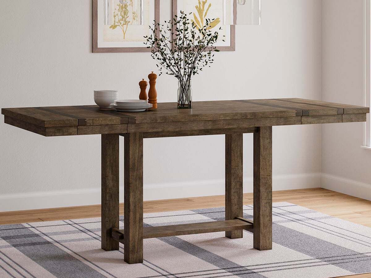 The Moriville RECT DRM Counter EXT Table, topped with a vase of branches, dishes, and salt and pepper shakers, stands on a plaid rug in a bright room.