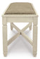 The Bolanburg Large UPH Dining Room Bench features a rectangular wooden frame, cross-braced legs in antiqued white, and a cushioned seat with beige heavy woven upholstery, shown from the front.