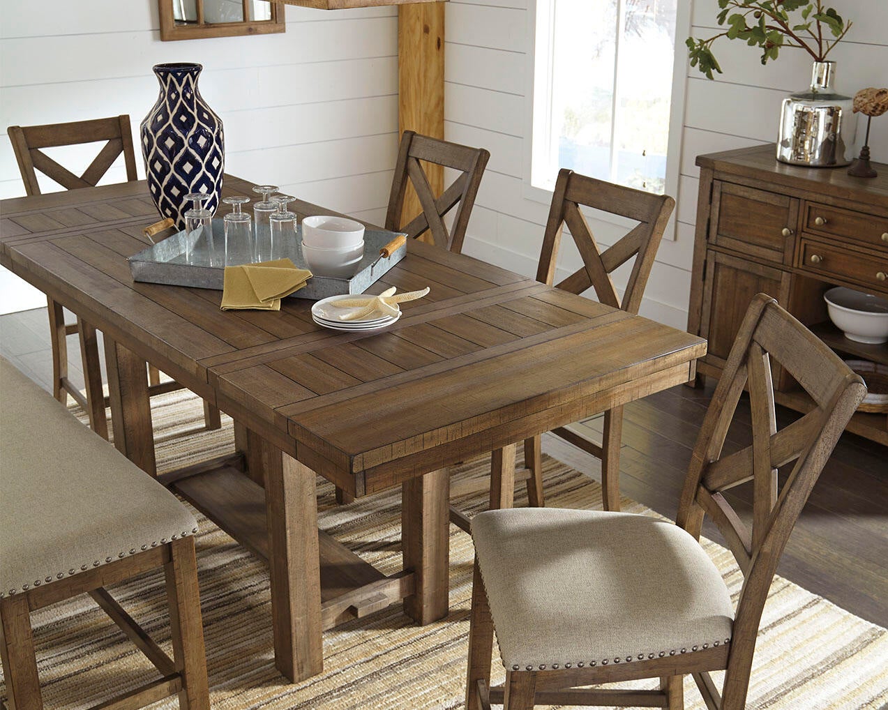 The Moriville RECT DRM Counter EXT Table, arranged with glasses, plates, silverware, and a vase, stands in a bright room with white walls and a rug. Matching chairs and a bench complete the loft-style dining set.