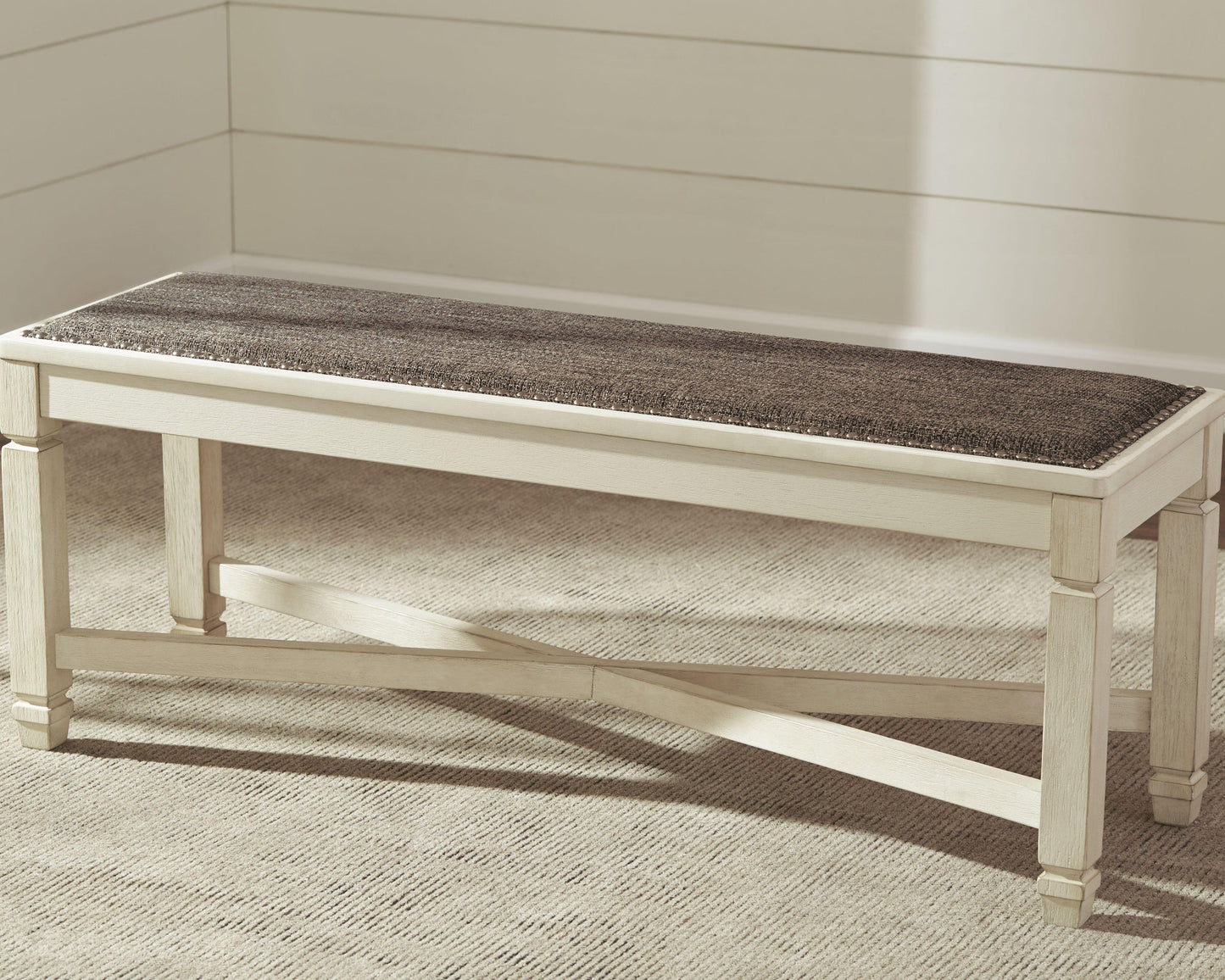 The Bolanburg Large UPH Dining Room Bench, featuring a dark cushioned seat in heavy woven fabric, is placed on a beige carpet against a white paneled wall.
