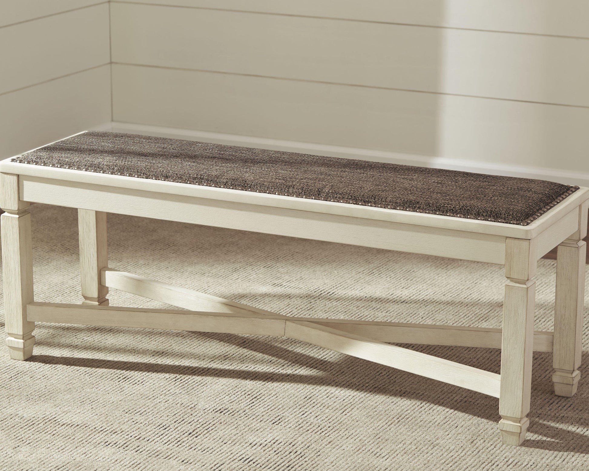 The Bolanburg Large UPH Dining Room Bench, featuring a dark cushioned seat in heavy woven fabric, is placed on a beige carpet against a white paneled wall.