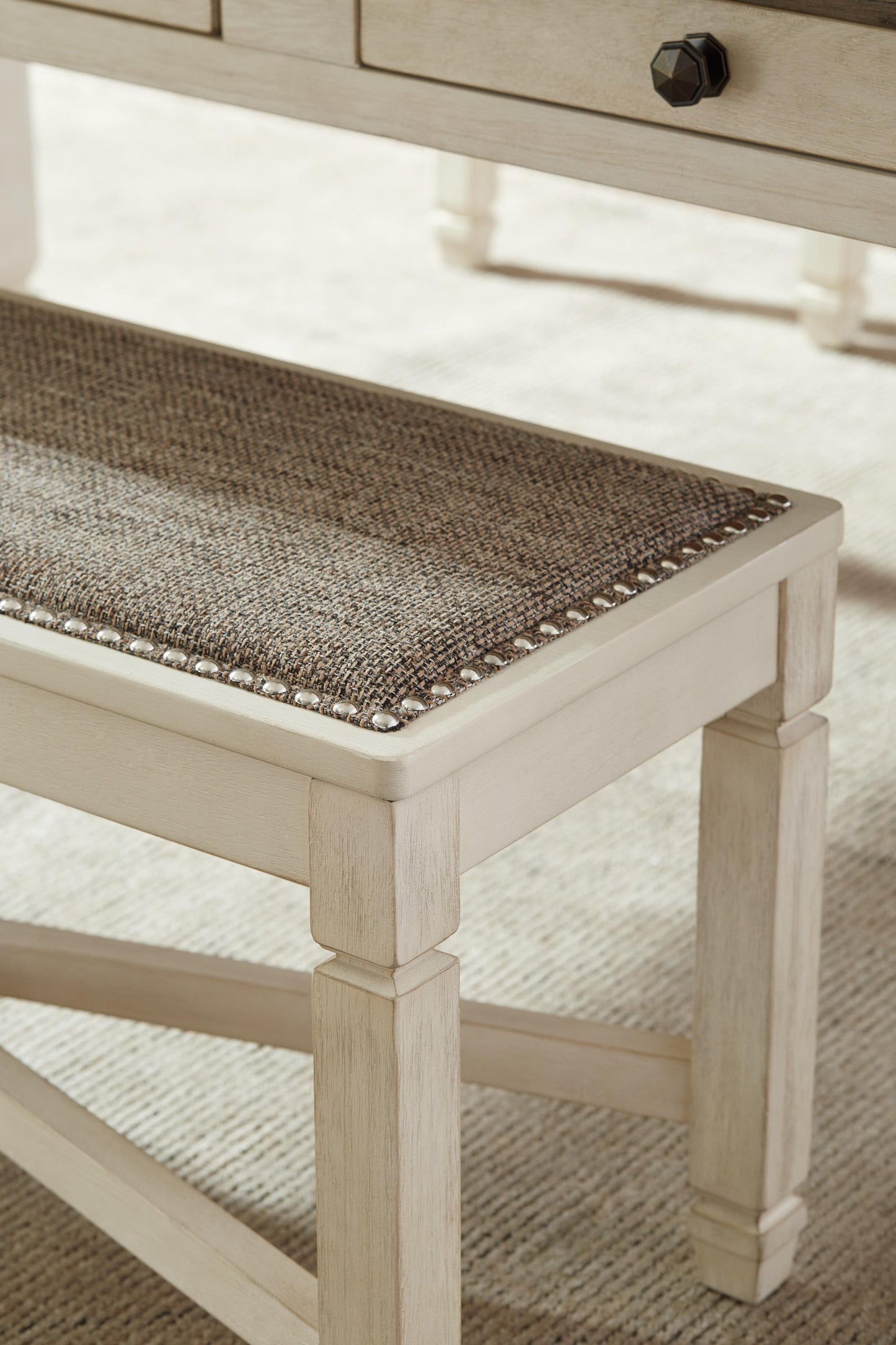 A close-up of the Bolanburg Large UPH Dining Room Bench with a heavy woven fabric seat and nailhead trim in antiqued white, set on a light-colored carpet next to a matching wooden table.