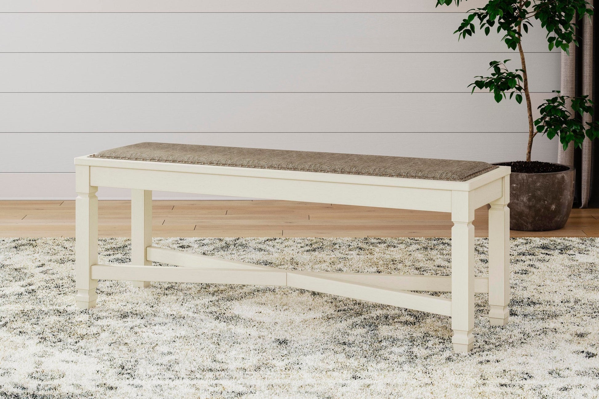 The Bolanburg Large UPH Dining Room Bench, featuring beige upholstery and antiqued white legs, is placed on a patterned rug in a room with light wood floors and a potted plant.