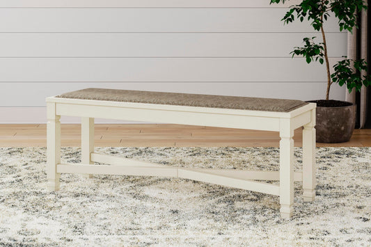 The Bolanburg Large UPH Dining Room Bench, featuring beige upholstery and antiqued white legs, is placed on a patterned rug in a room with light wood floors and a potted plant.