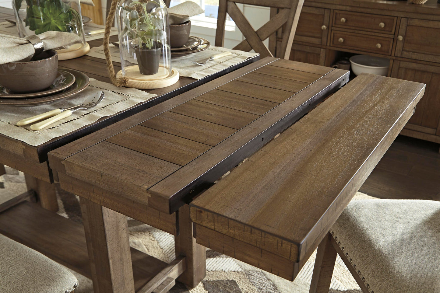 The Moriville RECT DRM Counter EXT Table, a wooden extendable dining table with its extension leaf partially pulled out, is set with dishes, utensils, and potted plants.
