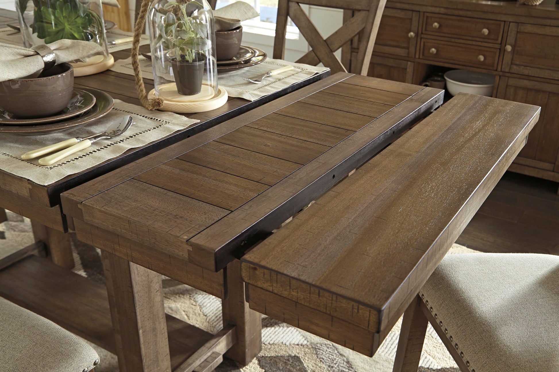 The Moriville RECT DRM Counter EXT Table, a wooden extendable dining table with its extension leaf partially pulled out, is set with dishes, utensils, and potted plants.