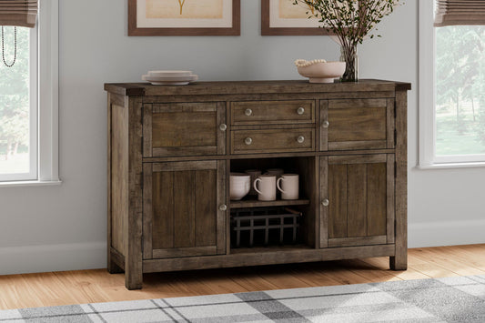 The Moriville Dining Room Server, with a distressed nutmeg finish and antiqued bronze-tone hardware, displays dishes and cups against the wall between two windows in a living room.