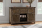 The Moriville Dining Room Server, with a distressed nutmeg finish and antiqued bronze-tone hardware, displays dishes and cups against the wall between two windows in a living room.