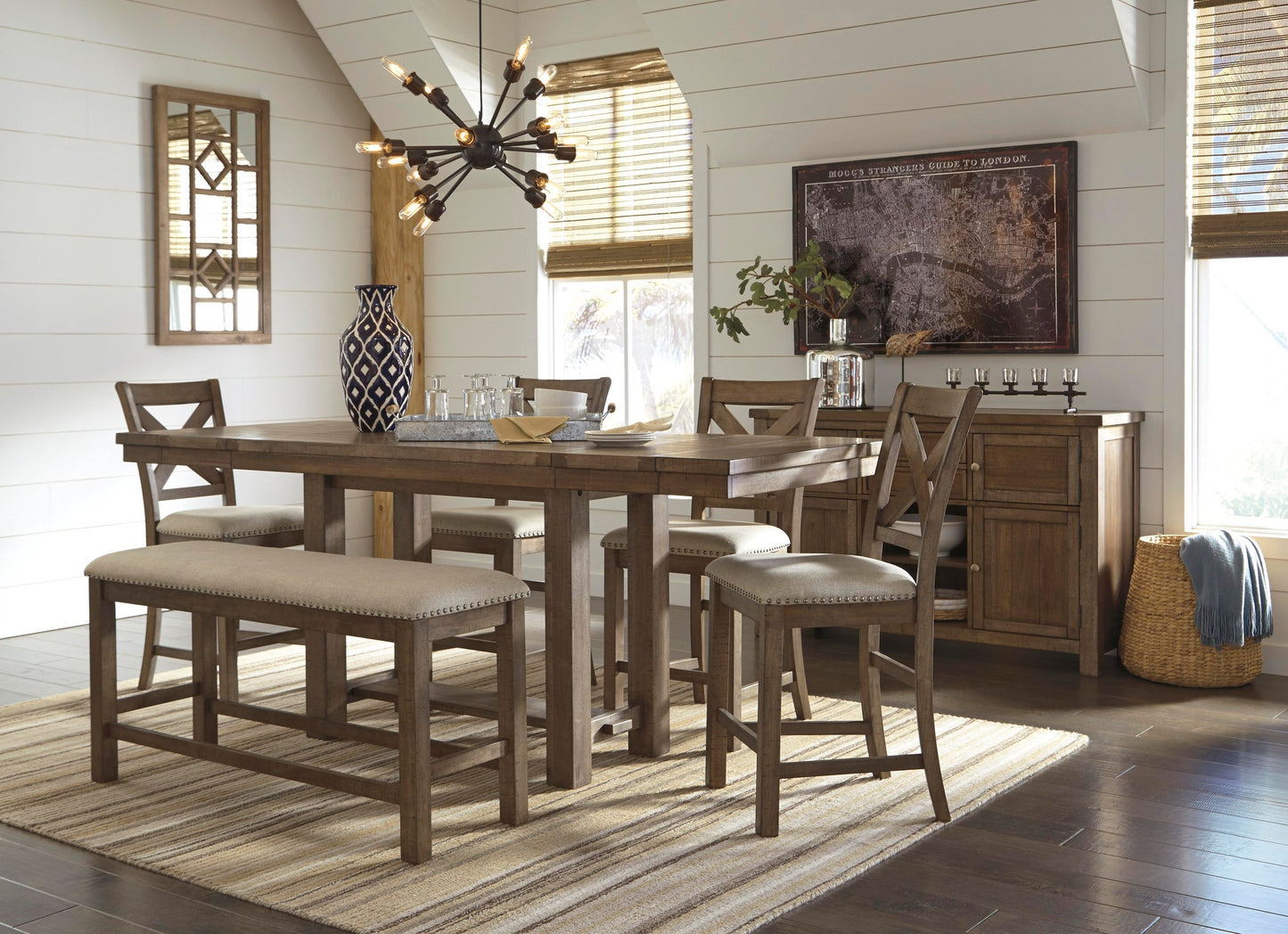 A modern dining room features the Moriville RECT DRM Counter EXT Table with matching chairs and bench in a distressed nutmeg finish on a striped rug, complemented by a contemporary chandelier, wall art, and natural light from large windows.