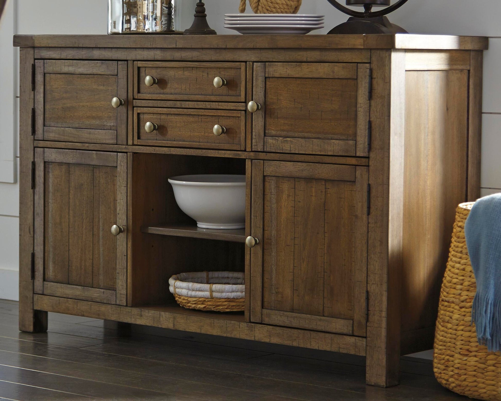 The Moriville Dining Room Server in a distressed nutmeg finish features four drawers, four cabinets with antiqued bronze-tone hardware, and an open shelf displaying a white bowl and folded towels on a dark wood floor.