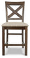 Moriville Upholstered Barstool (2/CN) features a wooden frame, "X" backrest, beige upholstered seat, brass nailhead trim, and a distressed nutmeg finish. Displayed from the front on a white background.
