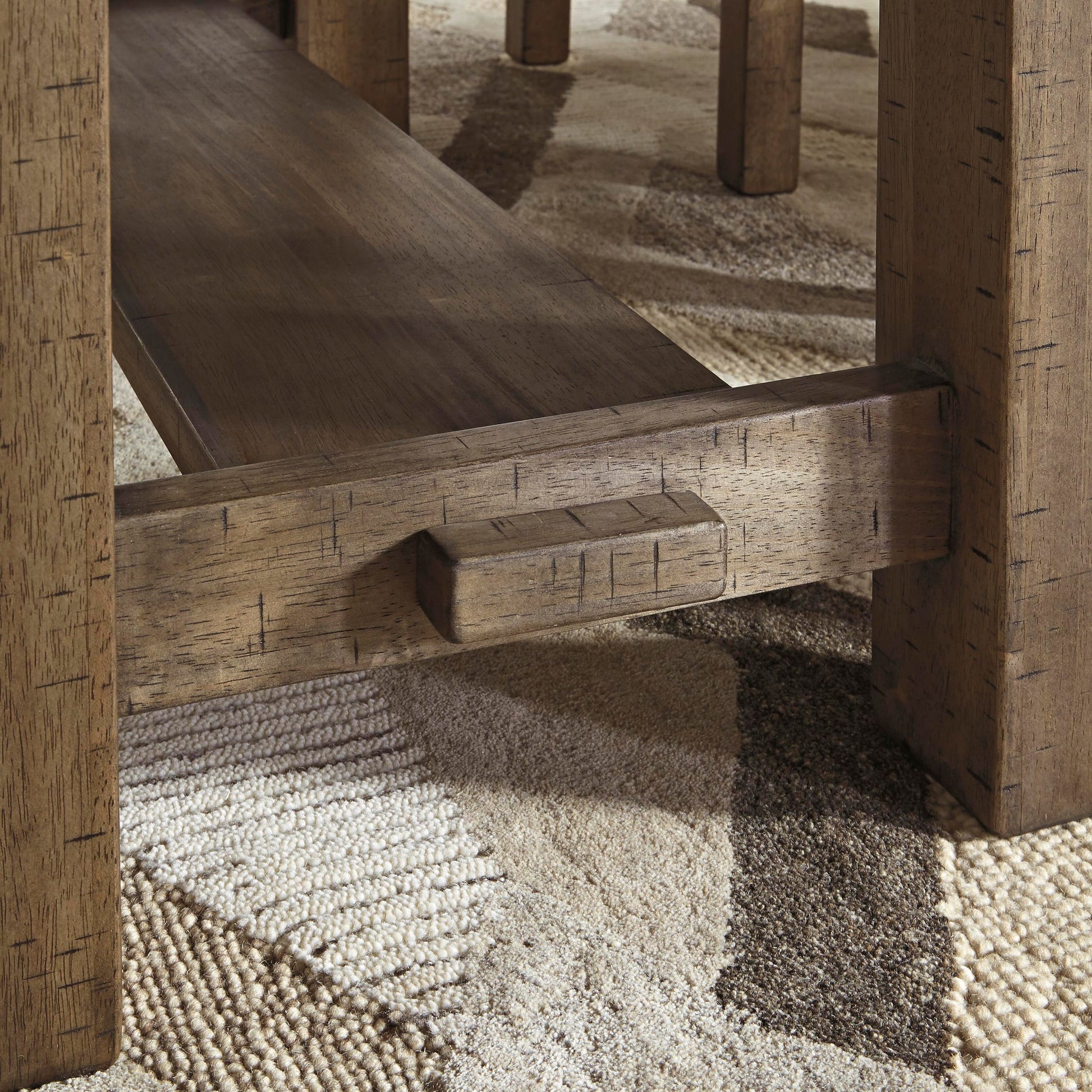 Close-up of the lower shelf and leg detail of the Moriville RECT DRM Counter EXT Table, featuring a distressed finish and shown on a patterned carpet.