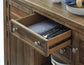 The Moriville Dining Room Server features a wooden kitchen drawer with antiqued bronze-tone hardware, opened to reveal neatly arranged silverware and a folded napkin. Plates and bowls are stacked on the countertop above.