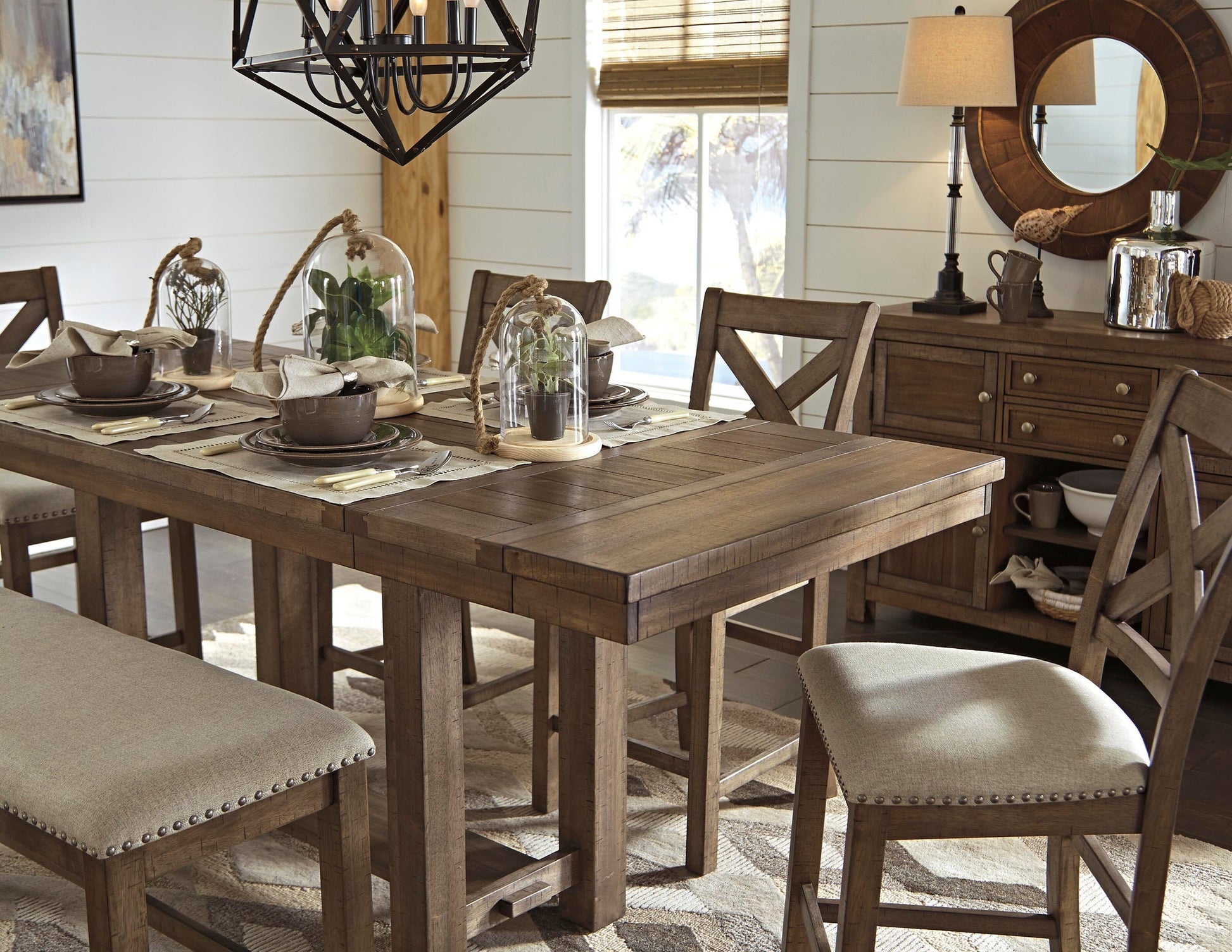 The Moriville RECT DRM Counter EXT Table in a distressed nutmeg finish seats six with beige-upholstered chairs, place settings, glass cloches with plants, and a sideboard topped by a round mirror in a bright dining room.