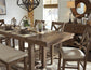 The Moriville RECT DRM Counter EXT Table in a distressed nutmeg finish seats six with beige-upholstered chairs, place settings, glass cloches with plants, and a sideboard topped by a round mirror in a bright dining room.