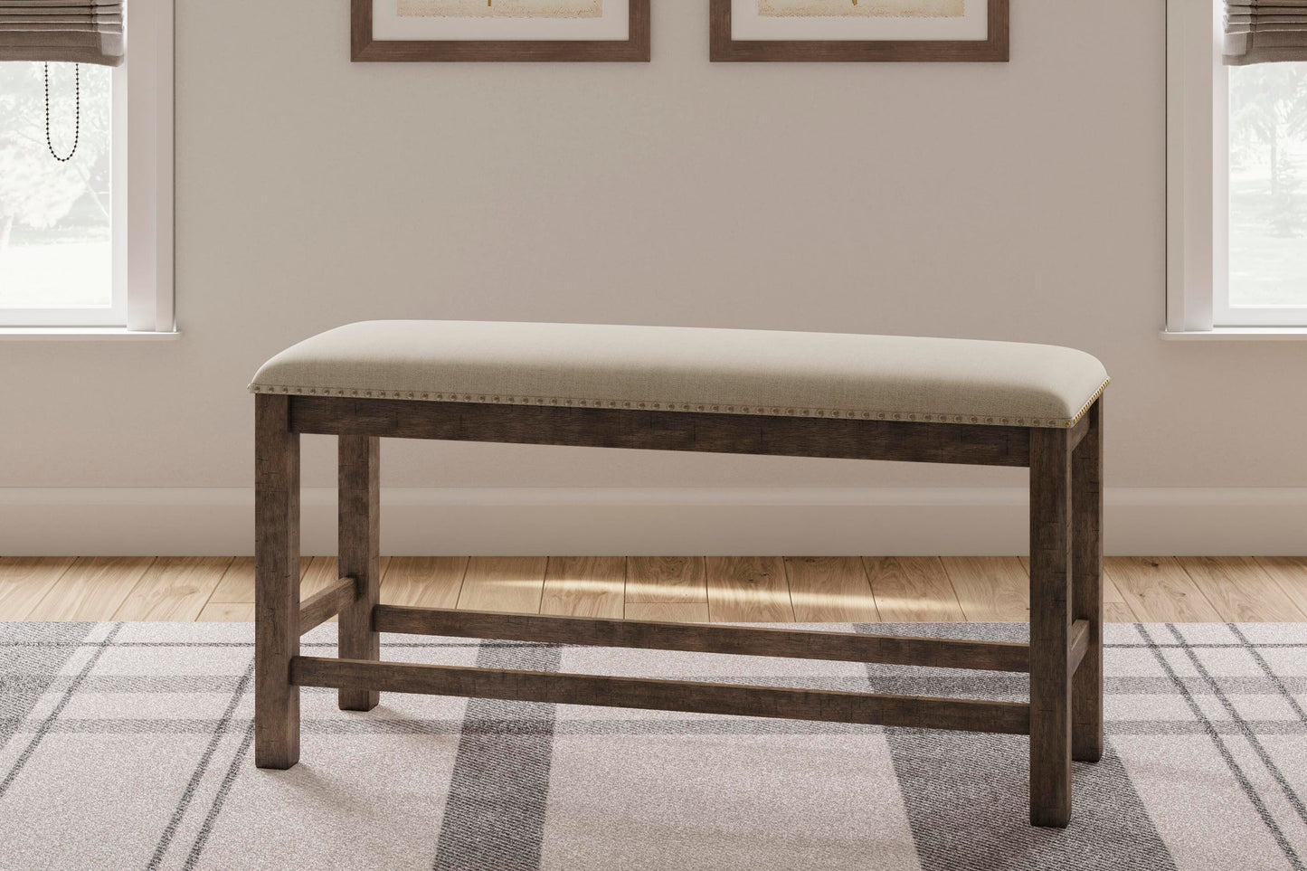 The Moriville Double UPH Bench, featuring a counter height and distressed nutmeg finish with a beige seat, sits on a patterned rug between two windows in a light-colored room.