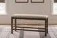 The Moriville Double UPH Bench, featuring a counter height and distressed nutmeg finish with a beige seat, sits on a patterned rug between two windows in a light-colored room.