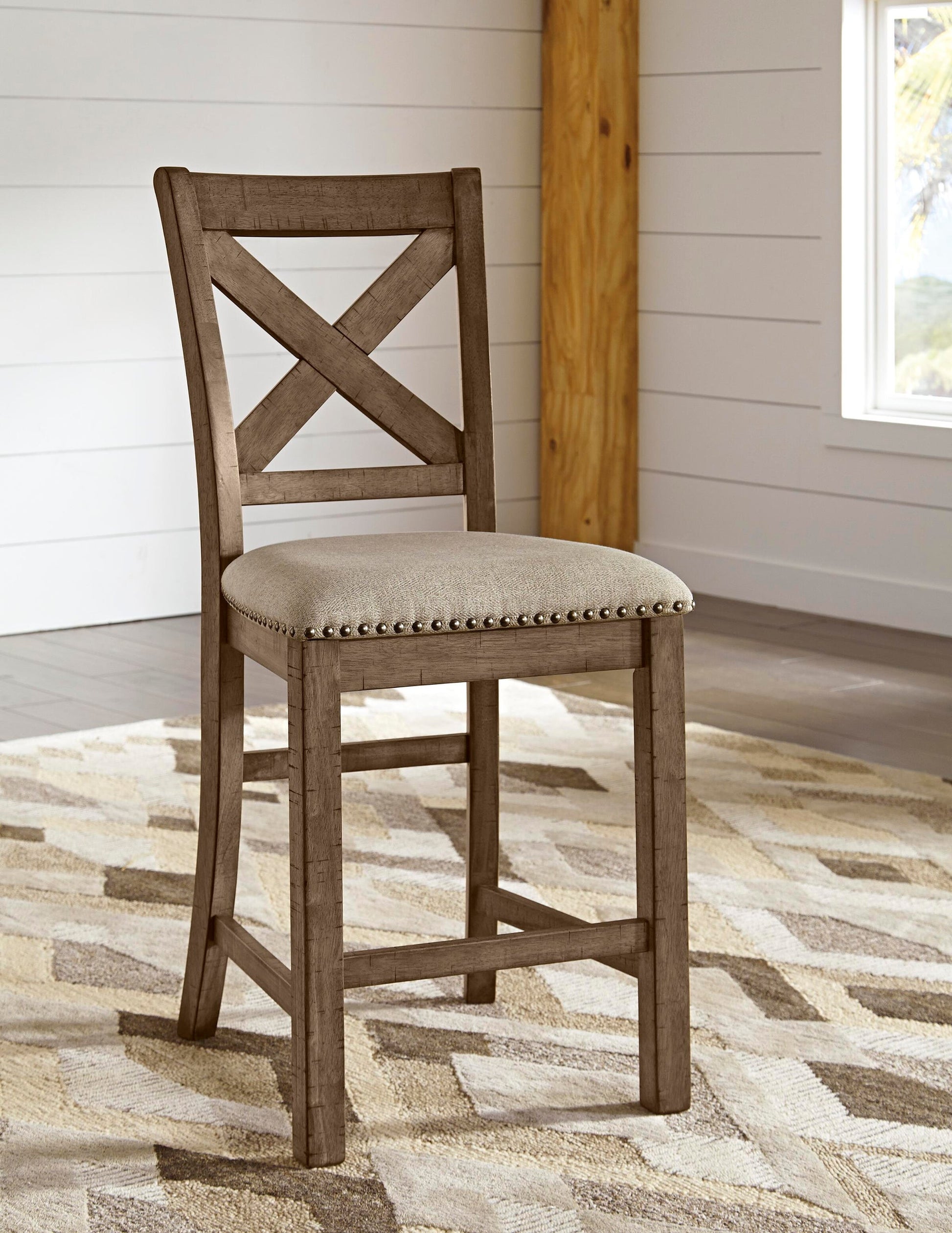 The Moriville Upholstered Barstool (2/CN) features a cross-back wooden design, cushioned beige fabric seats, and a distressed nutmeg finish—perfect for rooms with white walls, wood accents, and patterned area rugs.