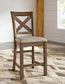 The Moriville Upholstered Barstool (2/CN) features a cross-back wooden design, cushioned beige fabric seats, and a distressed nutmeg finish—perfect for rooms with white walls, wood accents, and patterned area rugs.