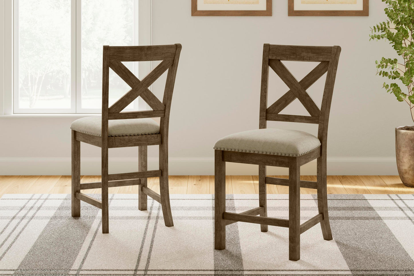 Two Moriville Upholstered Barstools with a distressed nutmeg finish and X-back design feature padded seats and are placed on a patterned rug in a bright room with a window and framed wall art.