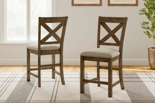 Two Moriville Upholstered Barstools with a distressed nutmeg finish and X-back design feature padded seats and are placed on a patterned rug in a bright room with a window and framed wall art.