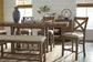 A wooden dining table with matching chairs and the Moriville Double UPH Bench sits on a striped rug, topped with a blue patterned vase, tray, and tableware. A sideboard with decorative items is in the background.