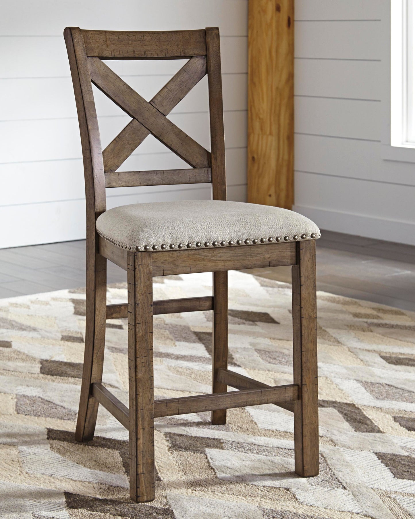 The Moriville Upholstered Barstool, with an X-back wooden frame and a light beige cushioned seat accented by nailhead trim, sits on a patterned rug in a room with white paneled walls.