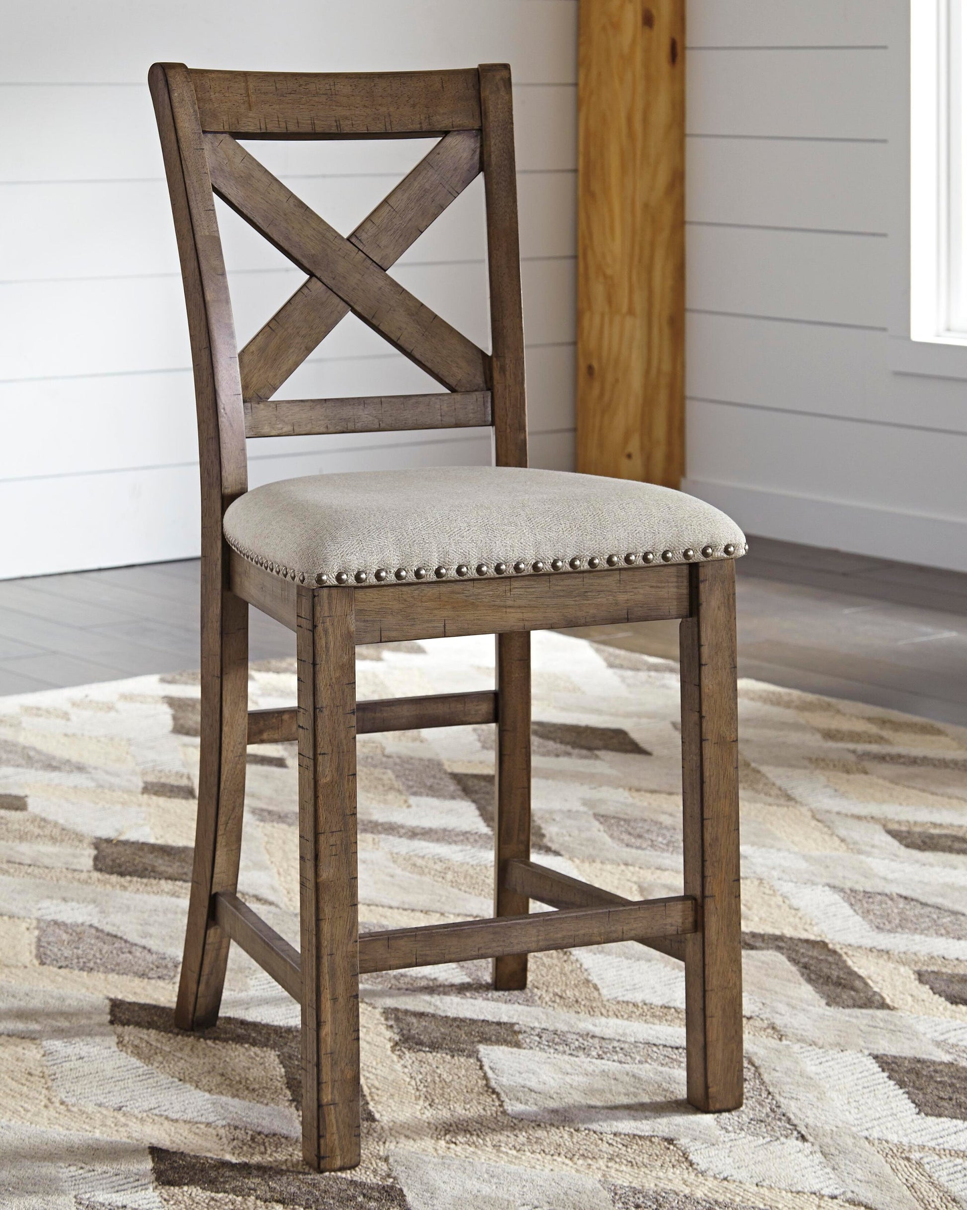 The Moriville Upholstered Barstool, with an X-back wooden frame and a light beige cushioned seat accented by nailhead trim, sits on a patterned rug in a room with white paneled walls.