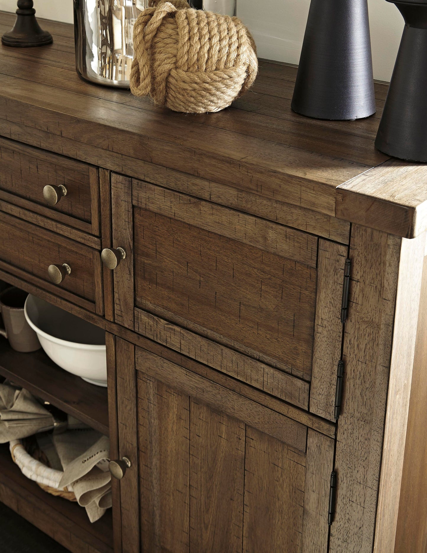 The Moriville Dining Room Server features drawers and cabinets in a distressed nutmeg finish, topped with a coiled rope, black vases, and a metallic container for stylish dining room storage.