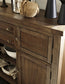 The Moriville Dining Room Server features drawers and cabinets in a distressed nutmeg finish, topped with a coiled rope, black vases, and a metallic container for stylish dining room storage.