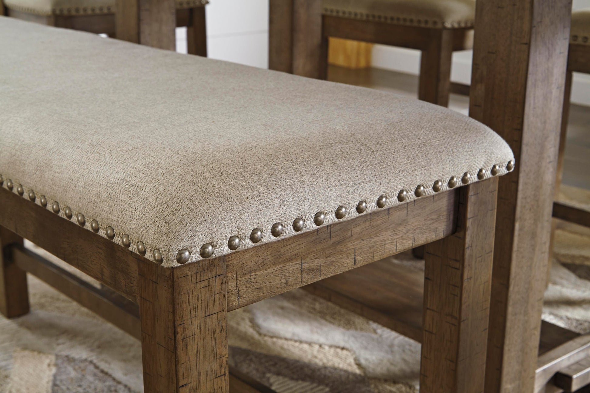 The Moriville Double UPH Bench (1/CN) features a light beige upholstered seat with antiqued nailhead trim and a distressed nutmeg finish, shown counter height next to matching wooden chairs.