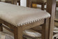 The Moriville Double UPH Bench (1/CN) features a light beige upholstered seat with antiqued nailhead trim and a distressed nutmeg finish, shown counter height next to matching wooden chairs.