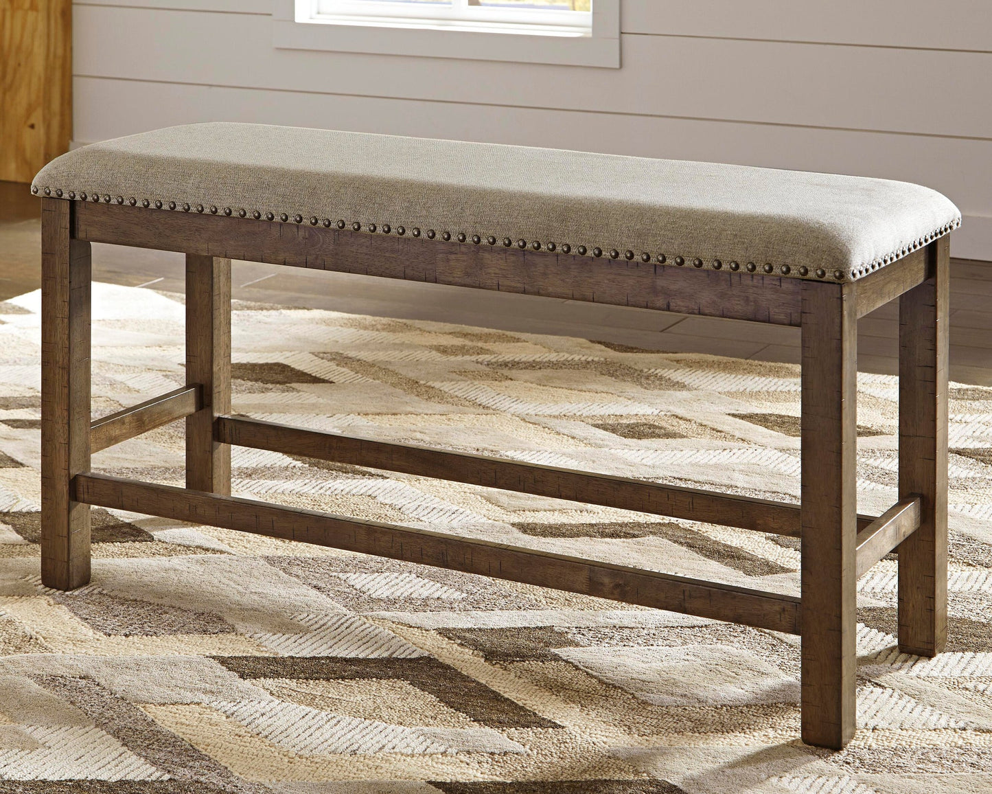 The Moriville Double UPH Bench, featuring a cushioned seat and antiqued nailhead trim, stands on a patterned rug in a bright room.